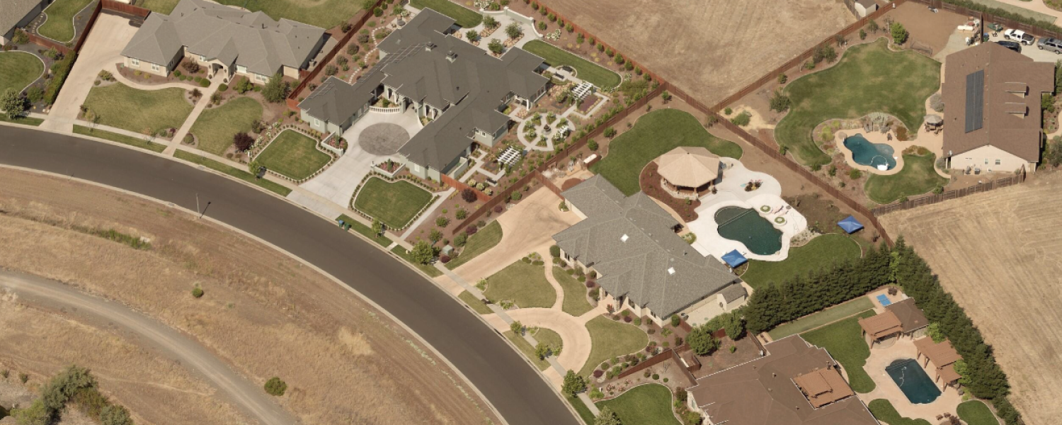 Aerial view of a suburban neighborhood featuring large homes with landscaped yards, circular driveways, and multiple swimming pools. The houses are separated by fences and surrounded by dry open fields and a curving paved road.