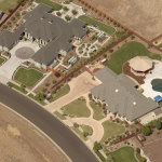 Aerial view of a suburban neighborhood featuring large homes with landscaped yards, circular driveways, and multiple swimming pools. The houses are separated by fences and surrounded by dry open fields and a curving paved road.