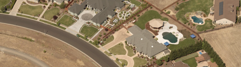 Aerial view of a suburban neighborhood featuring large homes with landscaped yards, circular driveways, and multiple swimming pools. The houses are separated by fences and surrounded by dry open fields and a curving paved road.