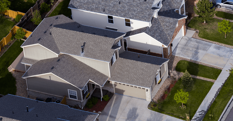 "Aerial view of a suburban neighborhood featuring two-story modern homes with gabled roofs, beige and gray exteriors, and neatly maintained front lawns and driveways. The houses are closely spaced, showcasing a planned residential community layout.