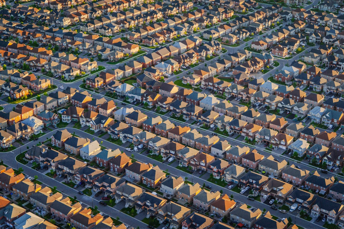 Aerial view of a densely packed suburban neighborhood with rows of similar single-family homes, arranged in a grid-like pattern with streets and driveways, taken in warm sunlight during late afternoon or early evening.