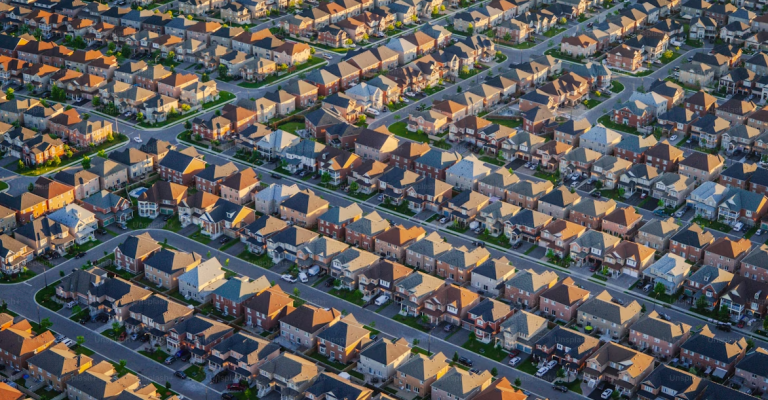 Aerial view of a densely packed suburban neighborhood with rows of similar single-family homes, arranged in a grid-like pattern with streets and driveways, taken in warm sunlight during late afternoon or early evening.