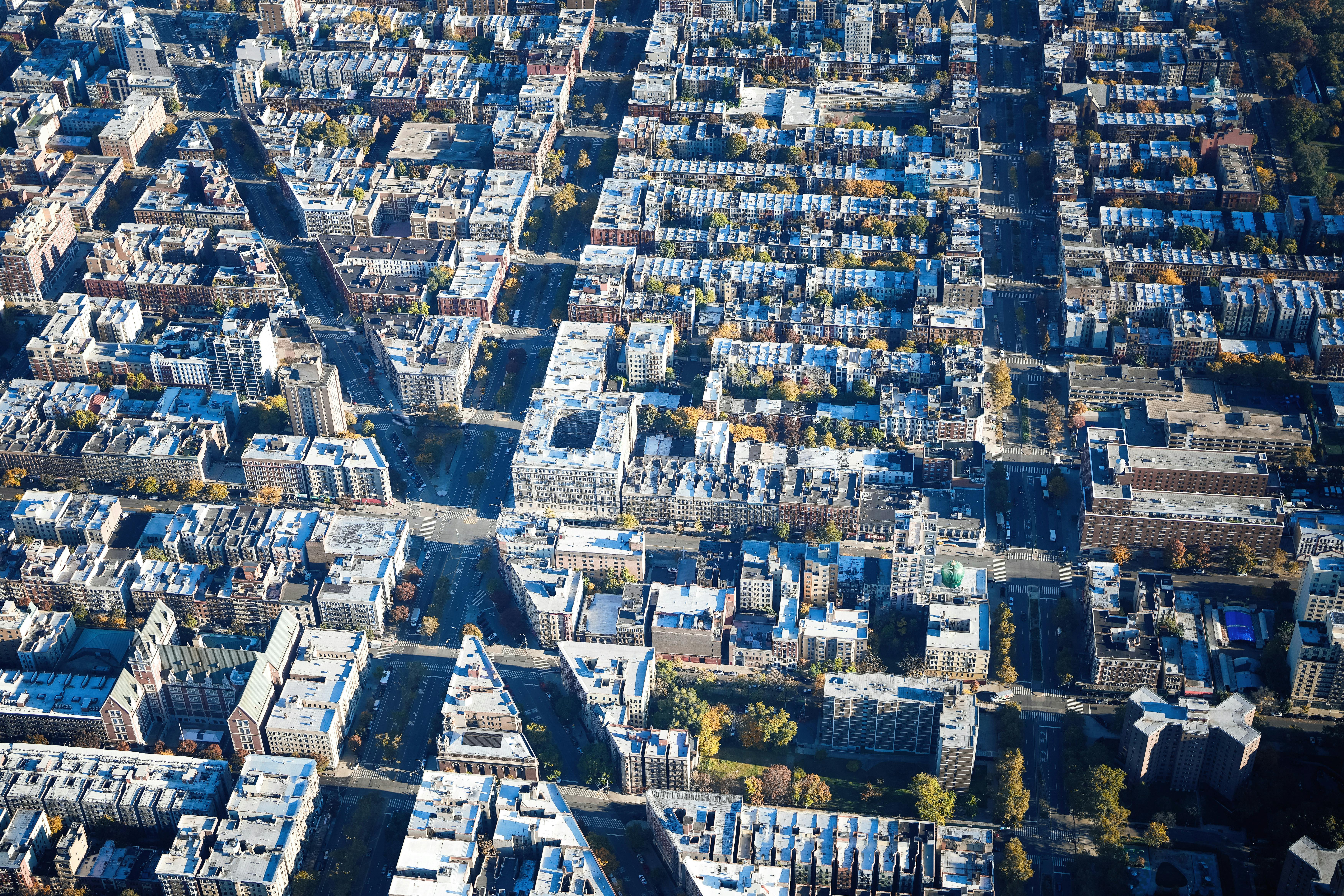 Aerial view of an urban city grid with densely packed apartment buildings and streets lined with trees. The buildings have mostly flat, light-colored rooftops, and the streets intersect at regular intervals, forming a structured grid pattern.