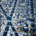 Aerial view of an urban city grid with densely packed apartment buildings and streets lined with trees. The buildings have mostly flat, light-colored rooftops, and the streets intersect at regular intervals, forming a structured grid pattern.