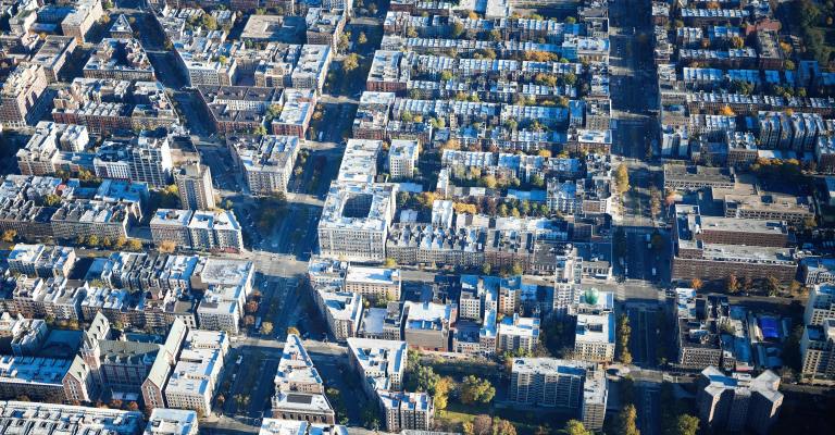 Aerial view of an urban city grid with densely packed apartment buildings and streets lined with trees. The buildings have mostly flat, light-colored rooftops, and the streets intersect at regular intervals, forming a structured grid pattern.